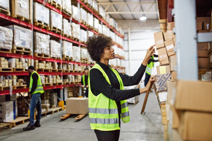 Female warehouse worker stacking boxes onto the rack. Woman working in large distribution warehouse. she is wearing uniform and reflective clothing, with colleagues working in background.
